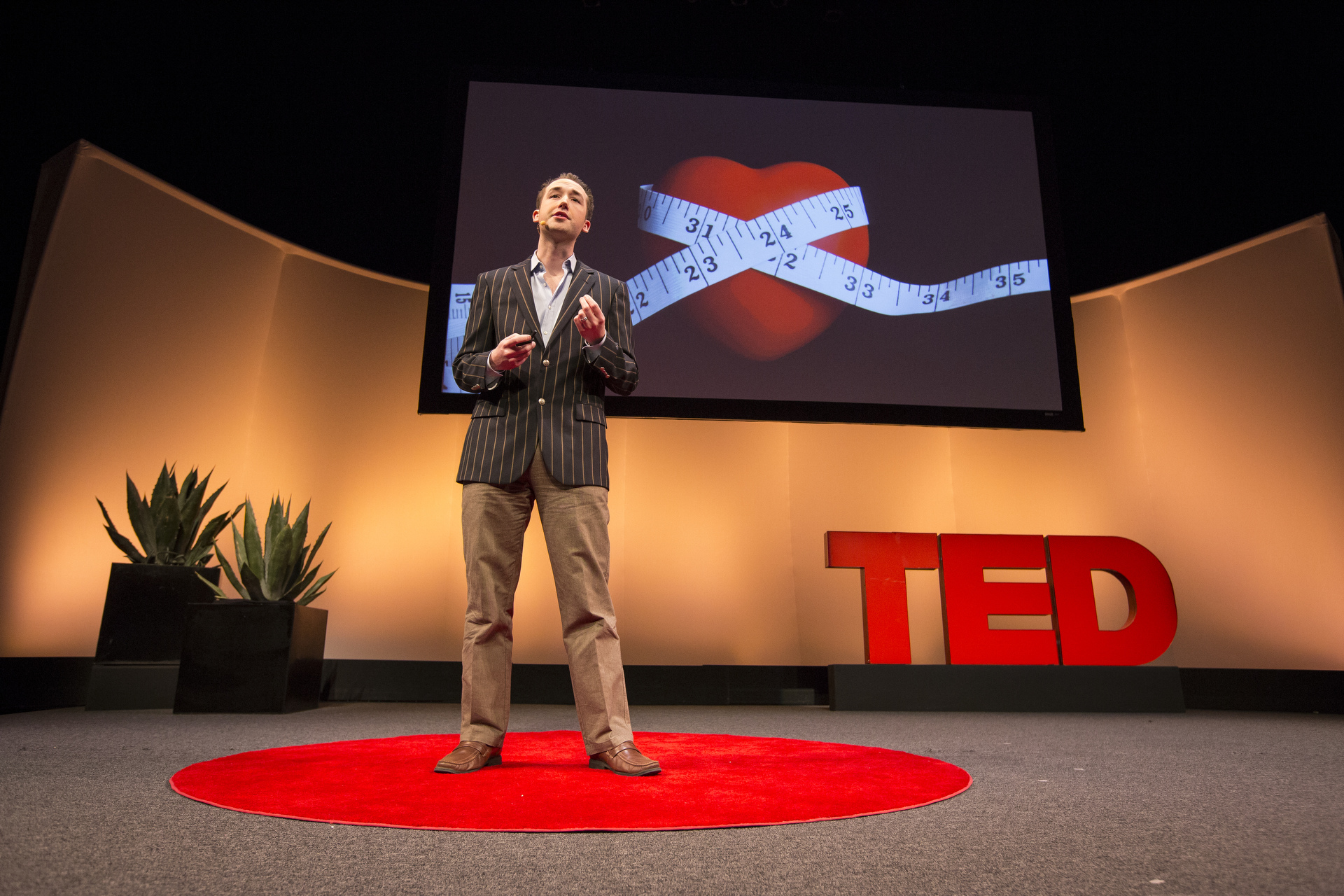 Paul Wicks speaking on the TED stage, a slide behind him showing a tape measure wrapped around a heart